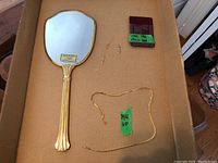 Full view of gold plated mirror, two gold pendants, and two gold chains arranged on brown cardboard.