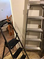 Photo showing both step ladders standing side by side indoors on a tiled floor near a wooden staircase and white wall.