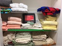 Image showing folded towels including dark and white colors on top shelf, various bedspreads folded in yellow, peach and cream colors, and a packaged king-size mattress protector in the middle shelf, with folded sheets and blankets on the bottom shelf.