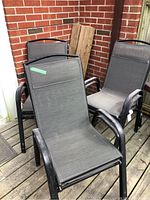 Set of six black metal patio chairs with mesh seats and backs, stacked and arranged on a wooden deck near a brick wall, visible dust and dirt on fabric surfaces.