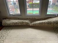 Wool rug partially rolled showing beige and green geometric pattern and standing on hardwood floor under large windows
