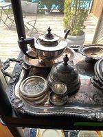 Silver plated serving pieces arranged on large ornate tray, showing tea pot with wooden handle, covered dishes, and cut glass coasters with silver rims.