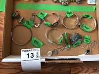 Top-down image of lot contents in a cardboard box showing the bracelets, ring, brooch, pin, and necklace with green price tags visible.