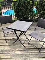 Photo of the folding patio table and two chairs set on a wooden deck outside near a pool, showing the entire set from a distance.