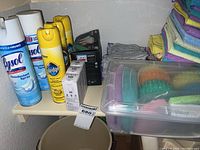 Shelf of Lysol and Pledge disinfectant spray cans, light bulb boxes, grey cleaning cloths, and a plastic container with cleaning sponges.
