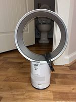 Front view showing the bladeless circular loop fan standing on a hardwood floor with power cord wrapped around the loop and a label sticker on the base.