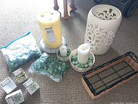 Fake candle lot including two white fake candles, green decorative rocks in bags and ceramic bowls, white ceramic lantern with tree cutout design, rectangular wicker basket, and three floral postcards.