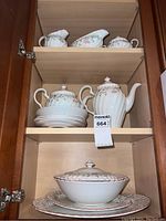 Photo of tea set inside wooden cabinet showing teapot, sugar bowl, creamer, covered serving bowl and plates stacked below.