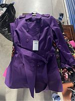 Photo showing a purple double-breasted trench coat with belt on top of a pile of jackets, including a floral pattern jacket underneath.