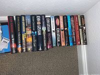 Side view of 20 John Grisham paperback books lined up on a white shelf showing titles and spines.