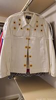 White ladies jacket with sunflower embroidery on chest and waist area, button front, hanging in closet.