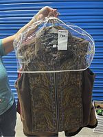 Photo of a brown ladies vest with multicolored embroidered pattern and faux fur collar on a hanger behind plastic cover.