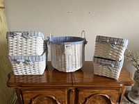 Five white wicker baskets arranged on wooden furniture, showing 1 tall round basket and four square baskets in two sizes with blue and white checkered liners