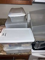 Several plastic storage containers with lids stacked on a countertop, one containing sewing supplies and another box holding furniture floor protectors.