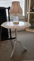 Full view of small cream-colored round accent table with carved floral designs and beige lamp on top, showing lamp cord and surroundings.