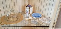 Whole lot displayed on shelf including brass tray, glass dome, plates, floral hat pin holder, small blue plates, lace doilies and decorative box.