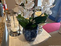 Blue ceramic vase holding white faux orchids next to glass candle holder and hourglass on white wooden surface.
