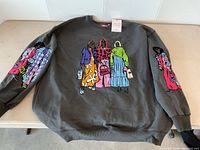 Front full view of the dark grey sweatshirt with colorful artwork of women on front and sleeves lying flat on a table.