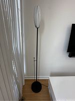 Full view of the tall black metal floor lamp with oval white and grey glass shade standing near a wall and curtain.