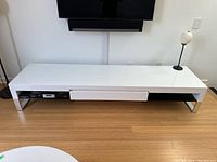 Front view of modern glossy white media console showing centre drawer and glass shelves underneath with aluminium legs.