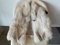 Front side of a light beige and off-white fox fur vest laid flat on a white chair, showing full fur texture and button closure.