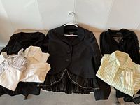 Photo of the full lot with all clothing items hung on hangers: black jacket, sheer polka dot blouse, ruffled black top, three cotton shirts in white and stripes.