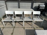 A frontal view of four white aluminum outdoor dining chairs with wooden armrests arranged side by side on a patio floor, showing the overall set and condition.