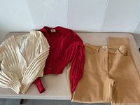 Camel-colored ultra suede pants with button and zipper closure, alongside two rib-knit cardigans, beige and red, laid on a light surface.