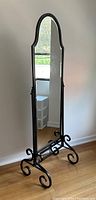 Front view of a full-length wrought iron cheval dressing mirror with arched top and black ornate scroll base, standing on a wood floor against a white wall.