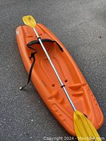 Orange kids kayak with black adjustable back rest and yellow paddle on pavement