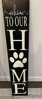Photo of tall black wooden porch sign with white lettering saying 'Welcome to Our Home' with a paw print replacing the letter 'O'.