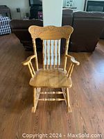 Front view of a wooden rocking chair with armrests and a curved top rail with carving, placed on hardwood flooring.