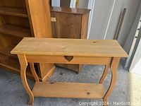 Wooden side table with curved legs and a bottom shelf, photographed in a storage area with other wooden furniture.