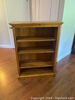Photo shows solid pine wooden bookshelf with four shelves, standing on a hardwood floor, in a well-lit room near doors and walls.
