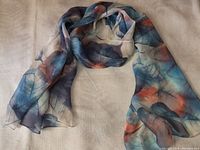 Lightweight scarf laid out displaying colorful abstract feather pattern in blue, orange, and gray on beige background.