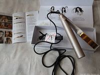 Hair curler with gold plates on light gold handle, showing embossed TYME logo, with cord and plug coiled, placed in original box alongside instruction leaflet with curling and styling diagrams.