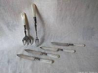 Photo of 7 vintage small cutlery pieces with mother of pearl handles on linen background, includes two forks and five knives or spreaders, showing the pearlescent handles and metal blades or tines.