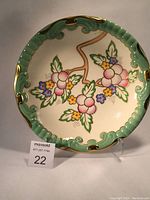 Top view of round ceramic wall plaque showing scalloped light green rim with gold accents and detailed hand-painted berries and flowers in pink, blue, yellow with green leaves.