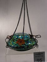 Photo shows the blue and green speckled glass top fill hummingbird feeder with black metal hanging chains and orange flower-shaped feeding ports.