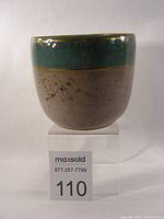 Front side view of a small ceramic bowl featuring turquoise blue upper rim and speckled beige lower body, placed on a clear stand with lot number tag visible.
