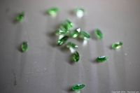 Clear photo showing sixteen small green oval-cut tourmalines scattered on a flat surface. Stones are visibly faceted and exhibit bright green color.