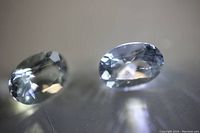 Two oval cut clear aquamarine gemstones displayed on a reflective surface, showing facets and transparency.