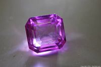 Close-up of bright pink sapphire gemstone showing Asscher cut on a flat surface, clear facets, and reflective properties.