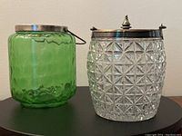 Pair of antique glass biscuit jars pictured side by side, one green with metal rim, the other clear glass with silver-plated lid and bail handle.