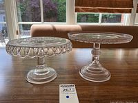 Two clear glass pedestal cake stands on wooden table, showing relative size and shape differences.