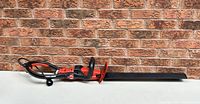 Full side view of Black & Decker electric hedge trimmer with blade cover, resting horizontally against a brick wall with the power cord extending from the handle.