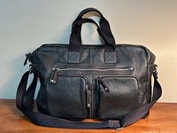 Front view of black leather messenger briefcase showing texture, handles, shoulder strap, and three zippered front pockets.