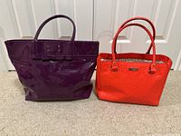 Front view of two Kate Spade tote bags, purple patent leather and orange textured, placed side by side on carpet.