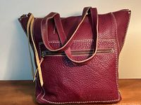 Front view of cranberry pebbled leather tote bag showing front zip pocket, contrast stitching and leather tassel pull.