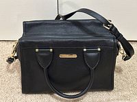 Front view of black pebbled leather satchel with top handles and detached strap resting behind